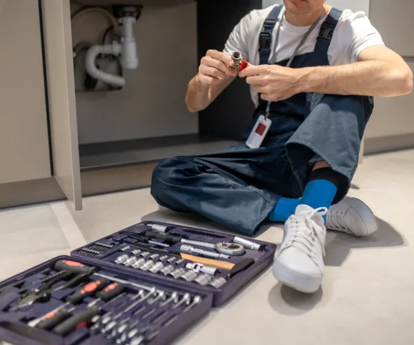 Right choice. Man in blue overalls looking at plumbing detail sitting on floor near open cabinet with sink and set of tools in kitchen, no face